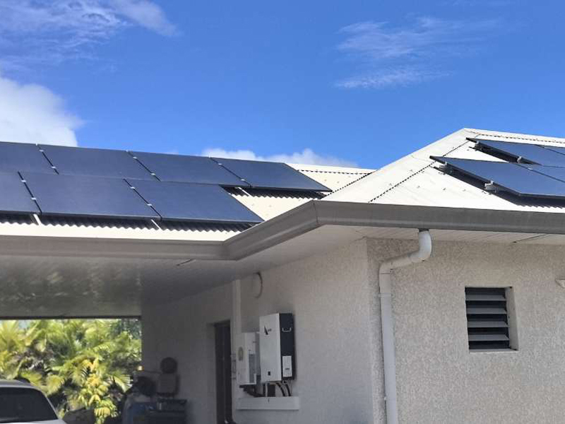 Multiple solar panels are installed on the roof of a light-colored house, set against a backdrop of clear blue skies and green vegetation.