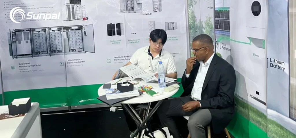 Sunpal representative reviewing brochures with a suited executive at a small table in front of BESS and PV product posters.