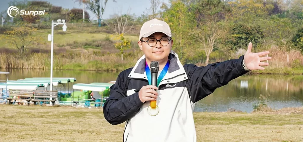 A Sunpal leader giving a speech outdoors during autumn team-building, holding a microphone and gesturing with one arm spread wide.