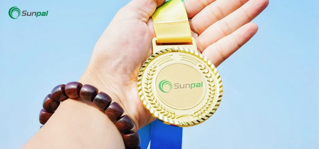 Close-up of a hand holding a gold medal with the Sunpal logo against a clear blue sky.