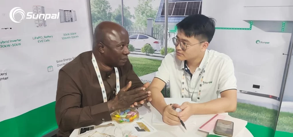 A Sunpal representative listens intently while discussing solar and battery solutions with a visitor at a small booth table.