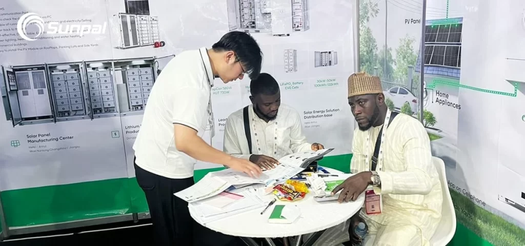 Sunpal representative reviews brochures with two visitors at a round table in front of BESS and PV system posters.